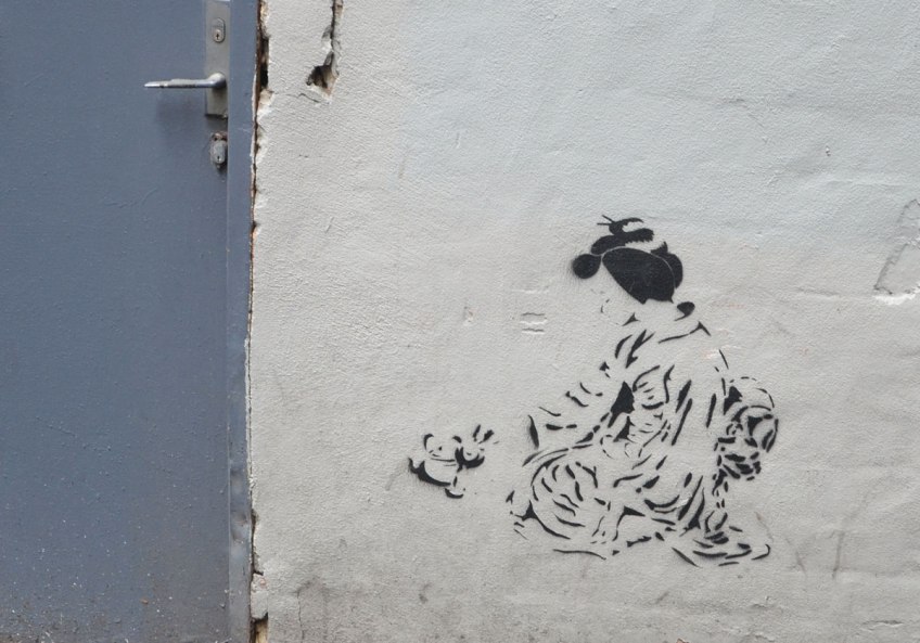 stencil graffiti of an Asian woman (Japanese?) crouching on the ground wearing a kimono, with her hair tied up. On a white wall beside a blue door