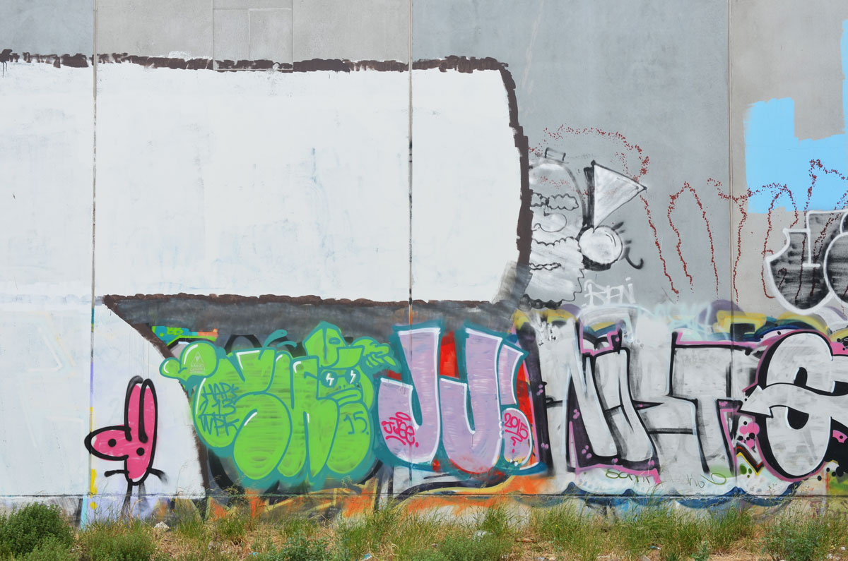 grey concrete wall with graffiti on it, large JJ in pink, a man's head, a green tag, railway tracks are in the foreground. 