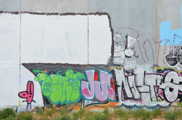 grey concrete wall with graffiti on it, large JJ in pink, a man's head, a green tag, railway tracks are in the foreground. 