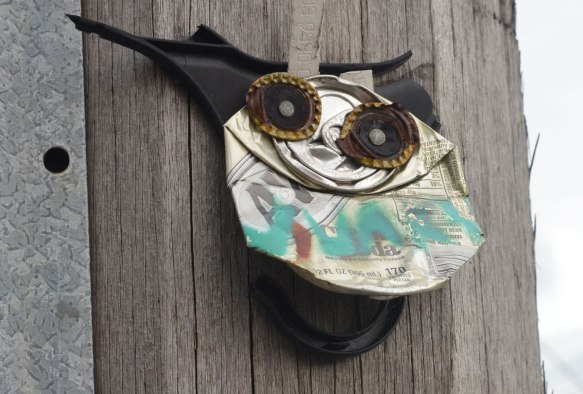 A junky creation nailed to a wooden power line pole with two bottle cap eyes and a broken black plastic hanger as a backbone