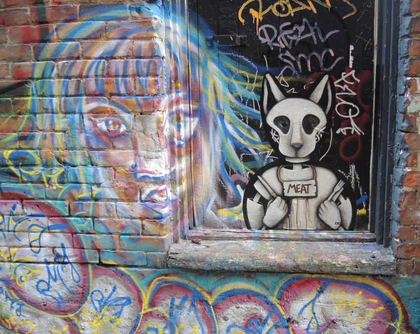 a poster of an creature wearing an animal mask sitting ina window sill, a knife in one hand and a fork in the other, looking like they are ready for dinner. A sign around its neck says 'meat'. A graffiti painting of a woman's face is beside the poster.