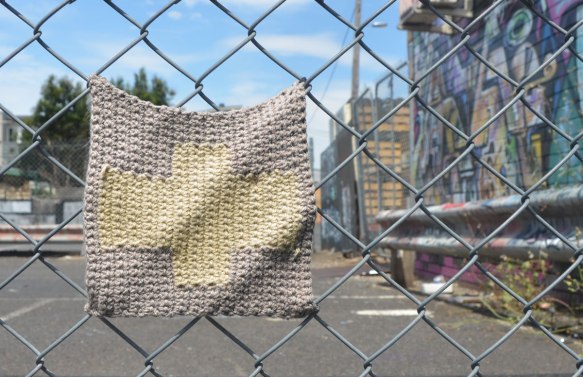 knitted square attached to a chain link fence, beige cross on grey background. Wall of graffiti out of focus in the background. 