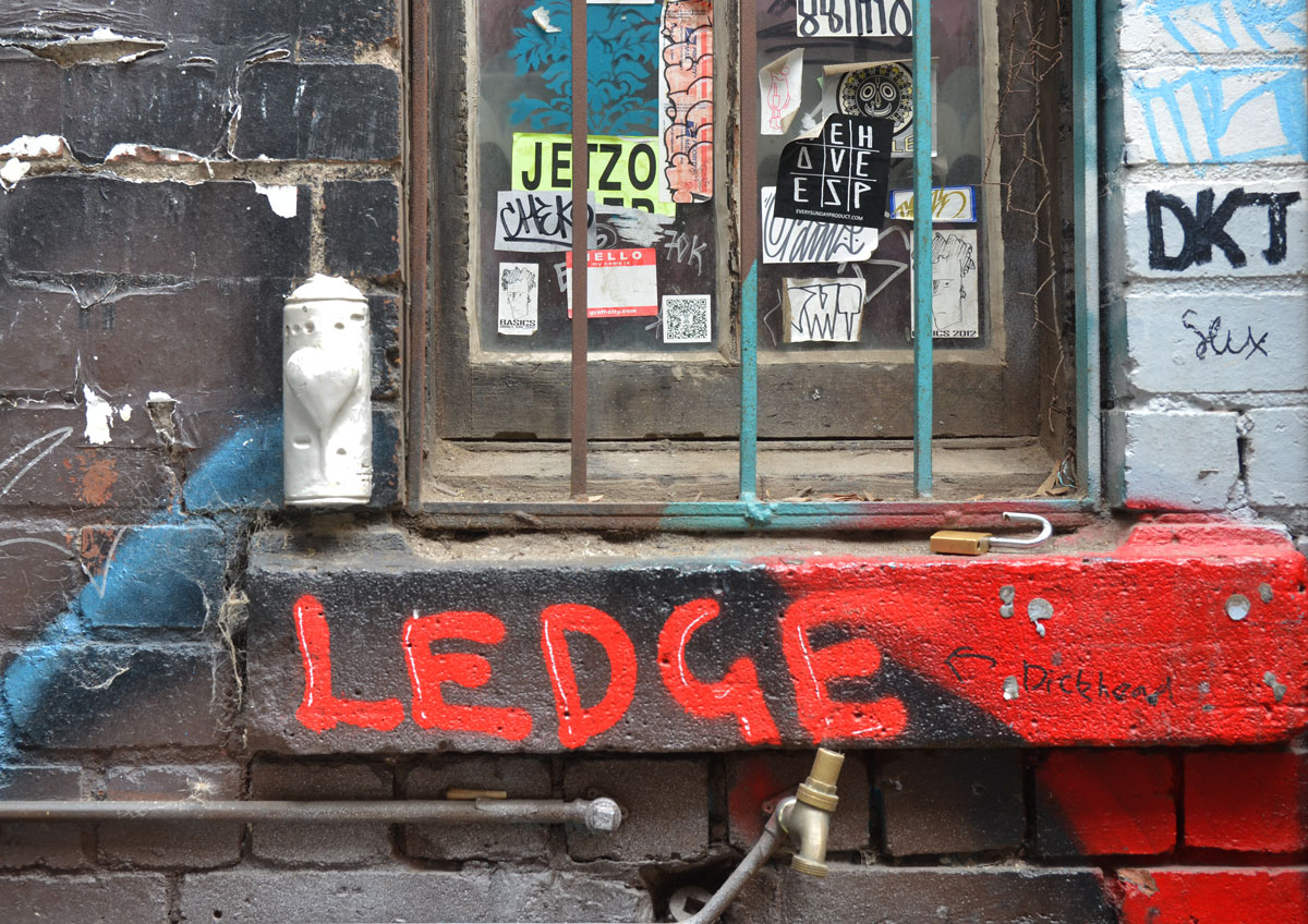 under a window in an alley. the window has metal bars. on the ledge under the window someone has spray painted in large red letters the word ledge