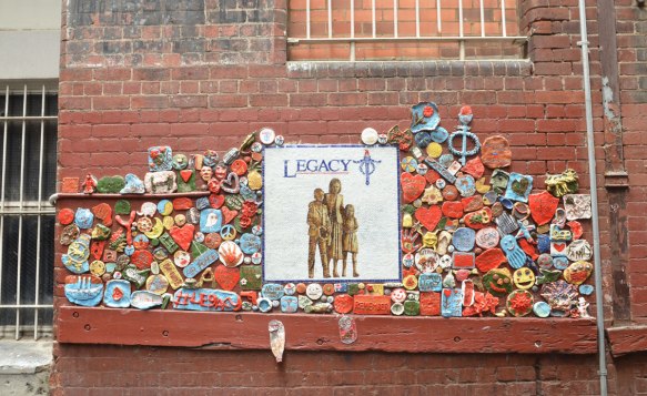 white square tile with the word legacy and a picture of three people, attached to an exterior wall with numerous small clay pieces around it.