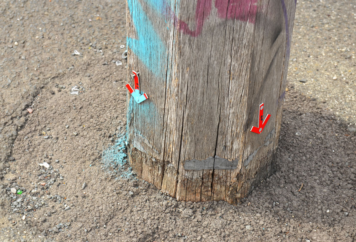 Two tiny little arrows attached to a telephone pole. Both of them point downwards. One is red and the other is red and turquoise. 