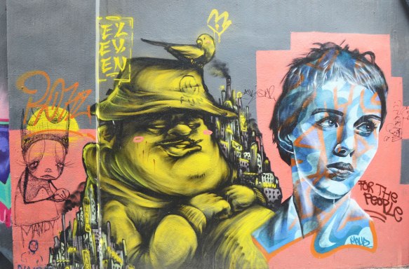 Three faces painting on a wall in an alley in Melbourne. On the right is a realistic painting of woman's head, in the middle is a caricature like picture of a man with a floppy hat and a bird on top of the hat. On the left is a black drawing of a sad looking girl on pink background. The word Mandy is written under the one in pink, and the words "For all the People" are written under the first woman. 