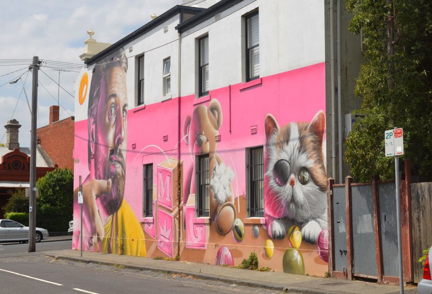 view of a mural by smug on Wood street in Melbourne, pink background, a man, a teddy bear and a kitten along with some wooden blocks and some marbles.