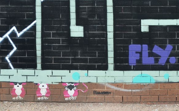 three small pink and white monkey wheatpastes under a large geometric (square) lettering tags. The monkeys are right by the ground. One is wearing sunglasses, one is wearing headphones and one is wearing a respirator. An interpretation of see no evil, hear no evil and speak no evil