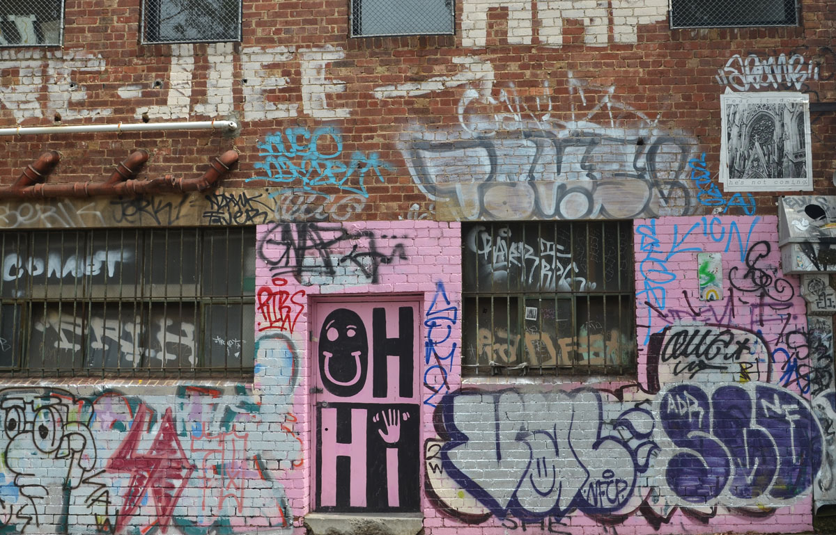 A lot of graffiti on a red brick wall. A door is in the middle and it has been painted pink. large black and pink letters are painted in it that say Oh Hi. The O in Oh is a happy face. The i in Hi is an arm and hand. 