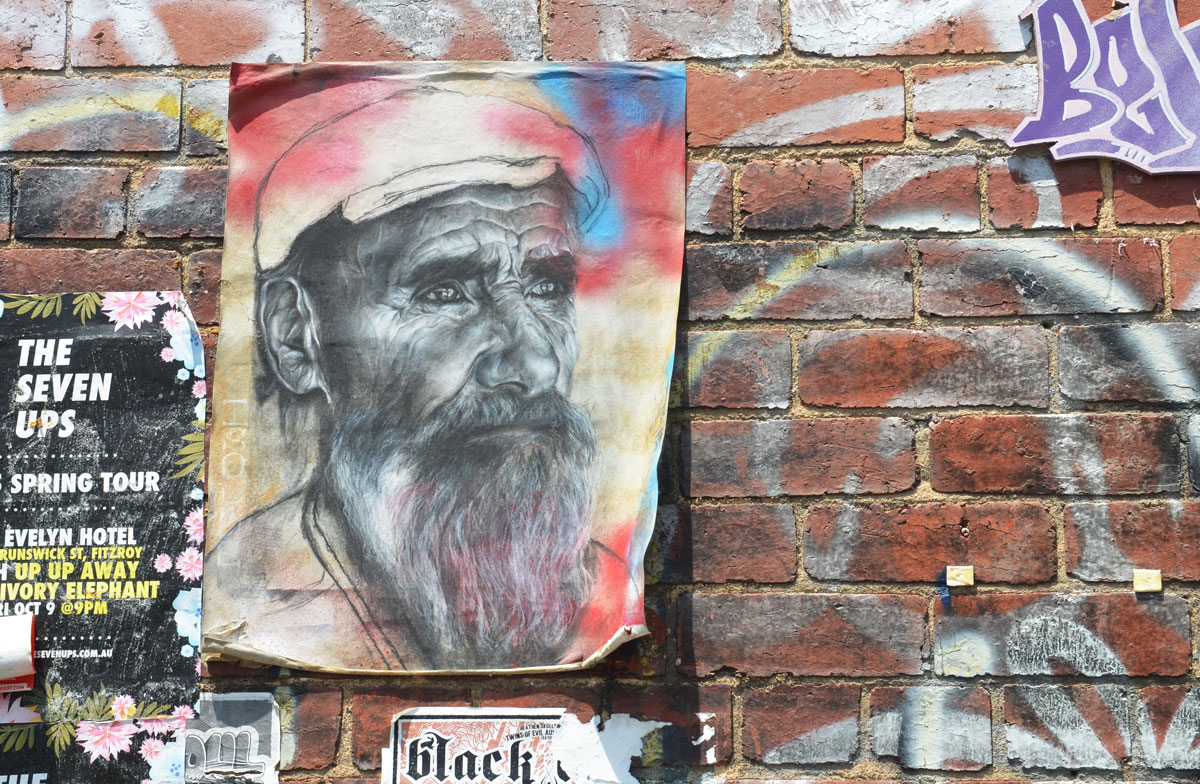 A drawing of an older man with a beard, wearing a white hat, on a piece of paper attached to a brick wall 
