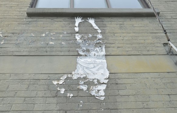 paper graffiti of a person hanging from a window sill (which is real), person is paper. It has started peeling away