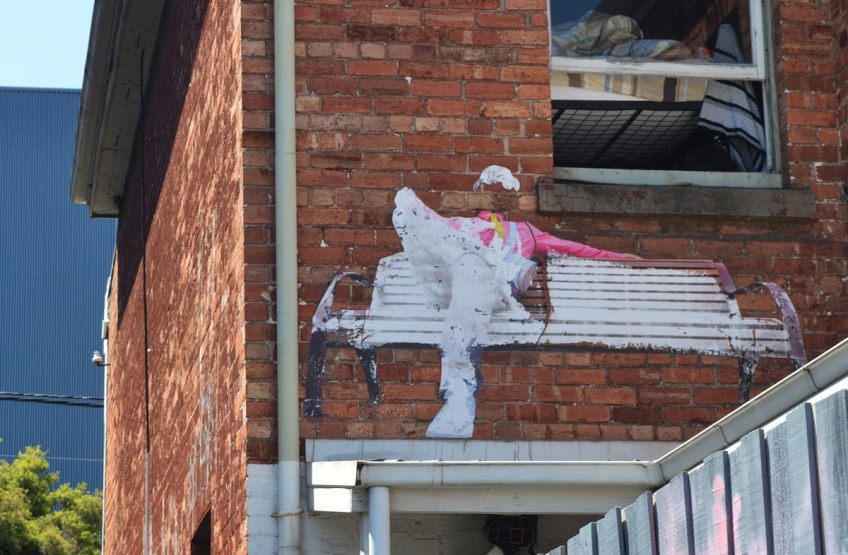 on a red brick wall, at second storey level, a large paste-up of a white wood bench with a person sitting on it. The person is wearing white trousers and shoes and a pink shirt.