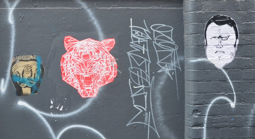 three paste ups, wheatpastes, on a black wall in an alley in central Melbourne. On the right is a man's head (black on white paper), in the middle is a pink tiger head with its mouth wide open and on the left is a head that looks like two heads merged together