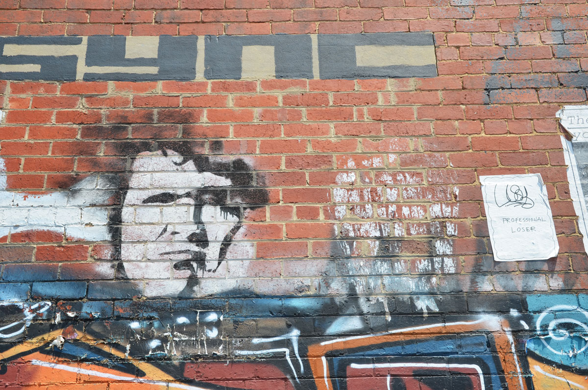graffiti on a brick wall, sync written in large block black letters, a black outlined white face, and a poster that says professional loser. 