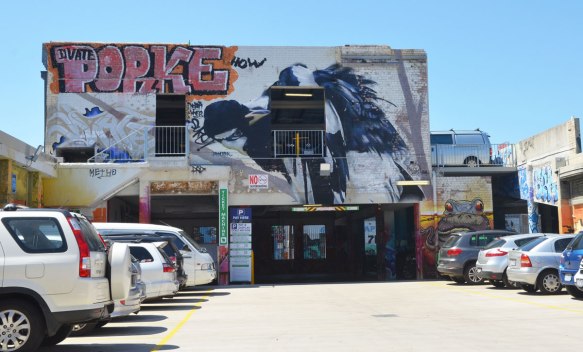 a car park with cars parked on either side, a wall (two storeys high) with a street art painting of a bird on it. 