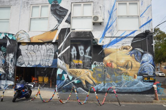 mural on the side of a two storey building of a woman lying on her side. A black bird is sitting on her shoulder. There is a yarn covered bike rack in front 