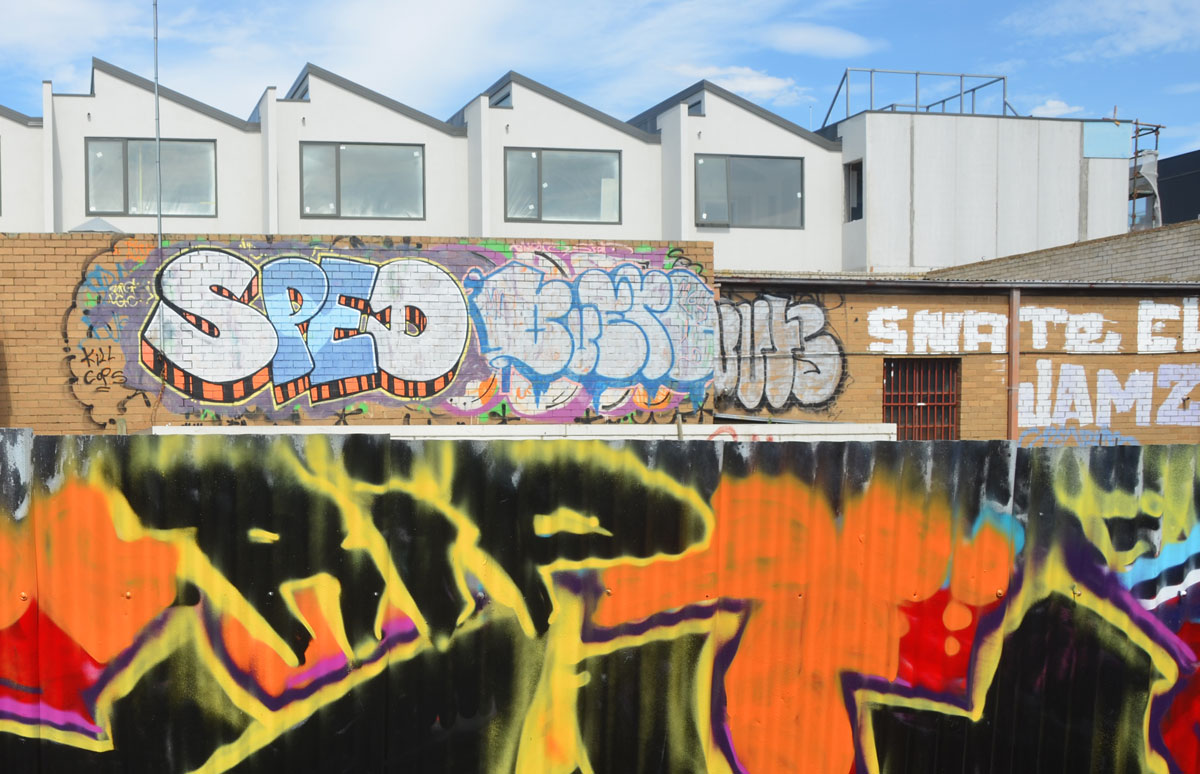 in the foreground is part of a corrugated metal fence with R I P in black that is part of a black, yellow, orange and red street art piece. In the background is a fence by some new houses. On the fence are a couple of tags, one of them is for SPED