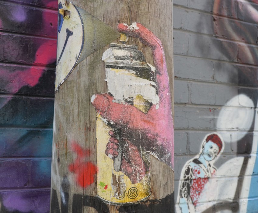 Two paste-ups on a pole, a pink hand on a spray paint can, aiming at a white happy face. Graffiti on walls in the background