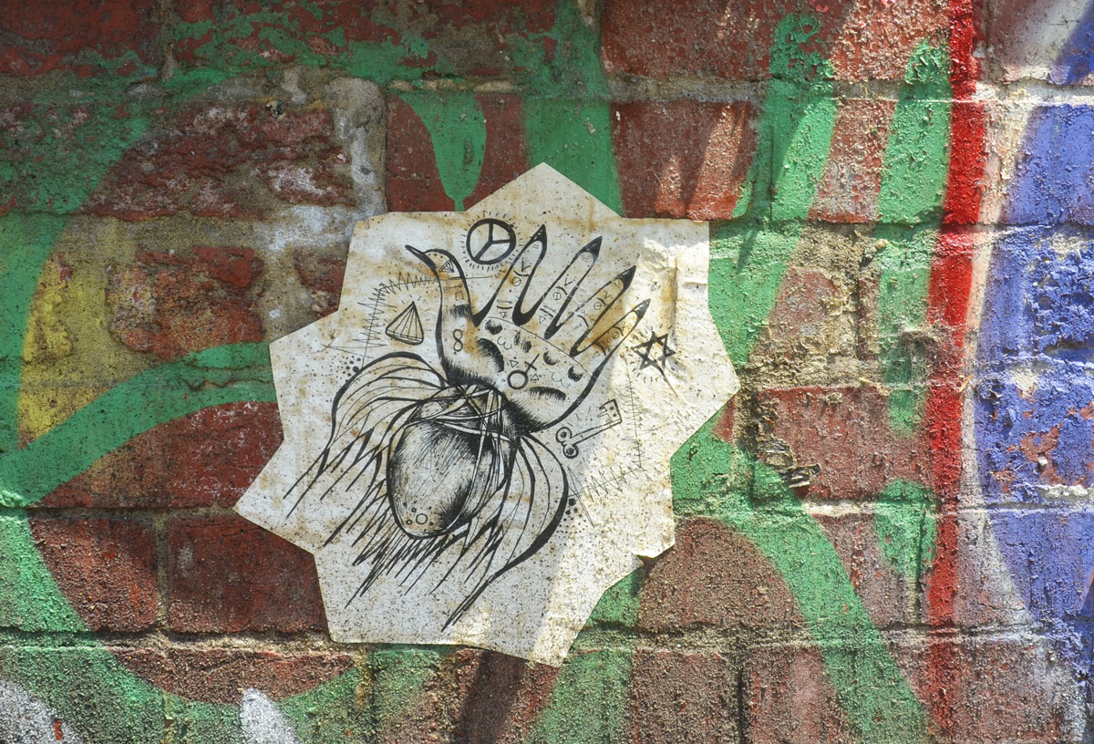 wheatpaste, black on white, graffiti on a brick wall that has some green spray paint on it, drawing is of a squid with a head that looks like a hand with five fingers. there are other symbols on it too, a peace sign, a key, a star of David, a diamond, the symbol that equals female