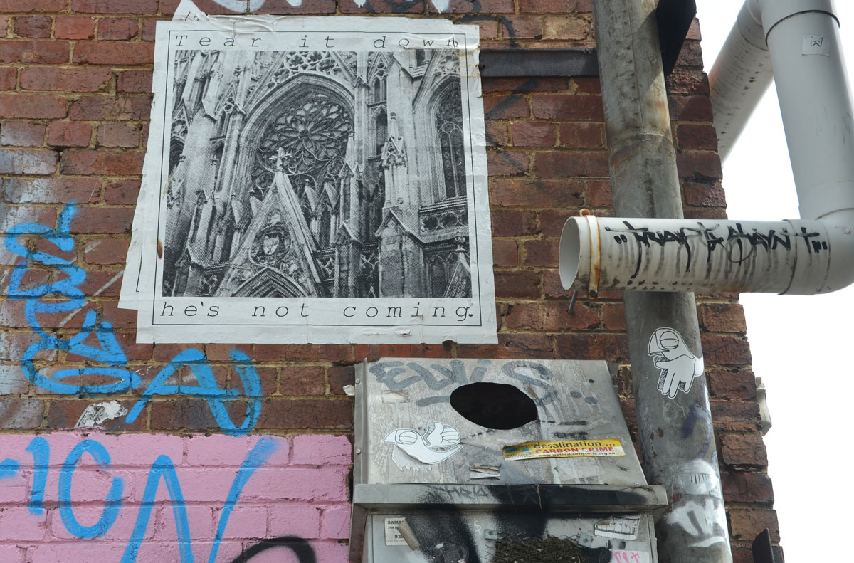poster graffiti high on a wall of a picture of a cathedral with the words "Tear it down. He's not coming"