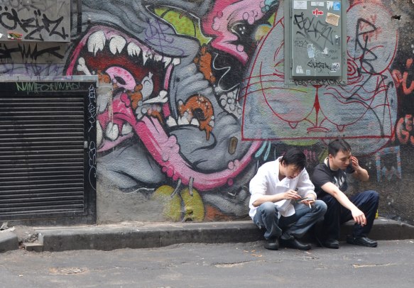 two young men sitting on a kerb smoking, they're sitting under a street art painting of a large mouth with a long tongue