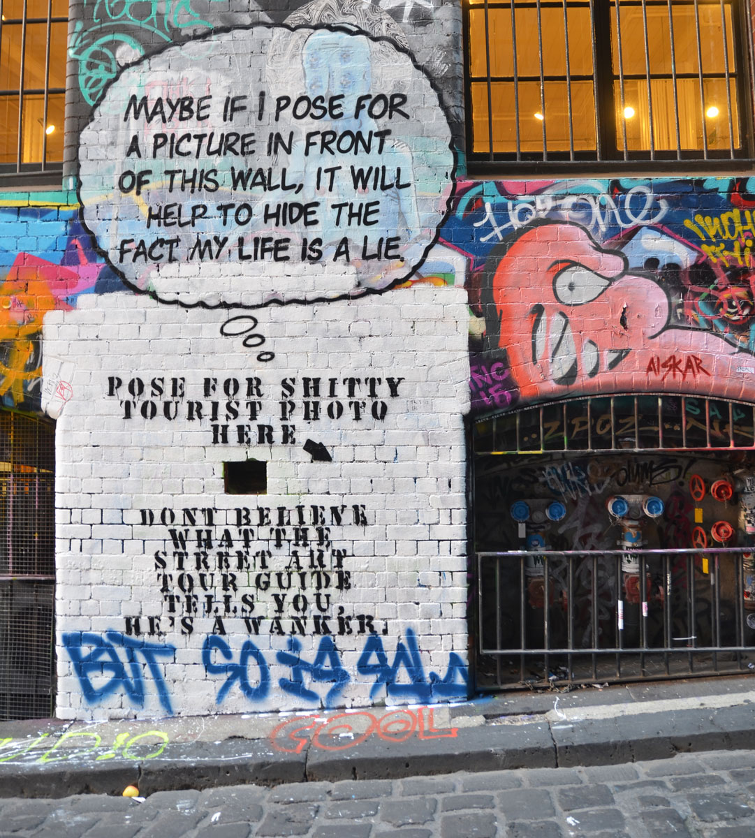 Lots of graffiti on the wall of Hosier Lane, windows with metal bars over them, including words that say "Maybe if I pose for a picture in front of this wall it will help to hide the fact that my life is a lie" Under those words, a large square section of the wall has been painted white and more words added: "Pose for shitty tourist photo here" with a black arrow pointing the place where you should stand. 