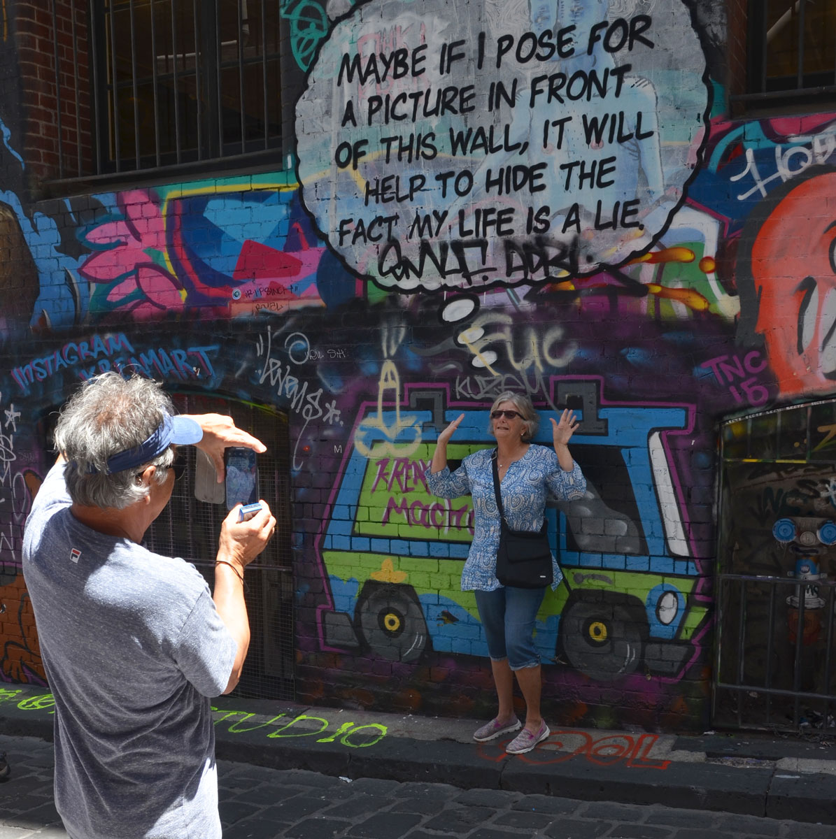Lots of graffiti on the wall of Hosier Lane, windows with metal bars over them, a woman poses in front of words that say "Maybe if I pose for a picture in front of this wall it will help to hide the fact that my life is a lie" while a man takes her picture with a smartphone camera
