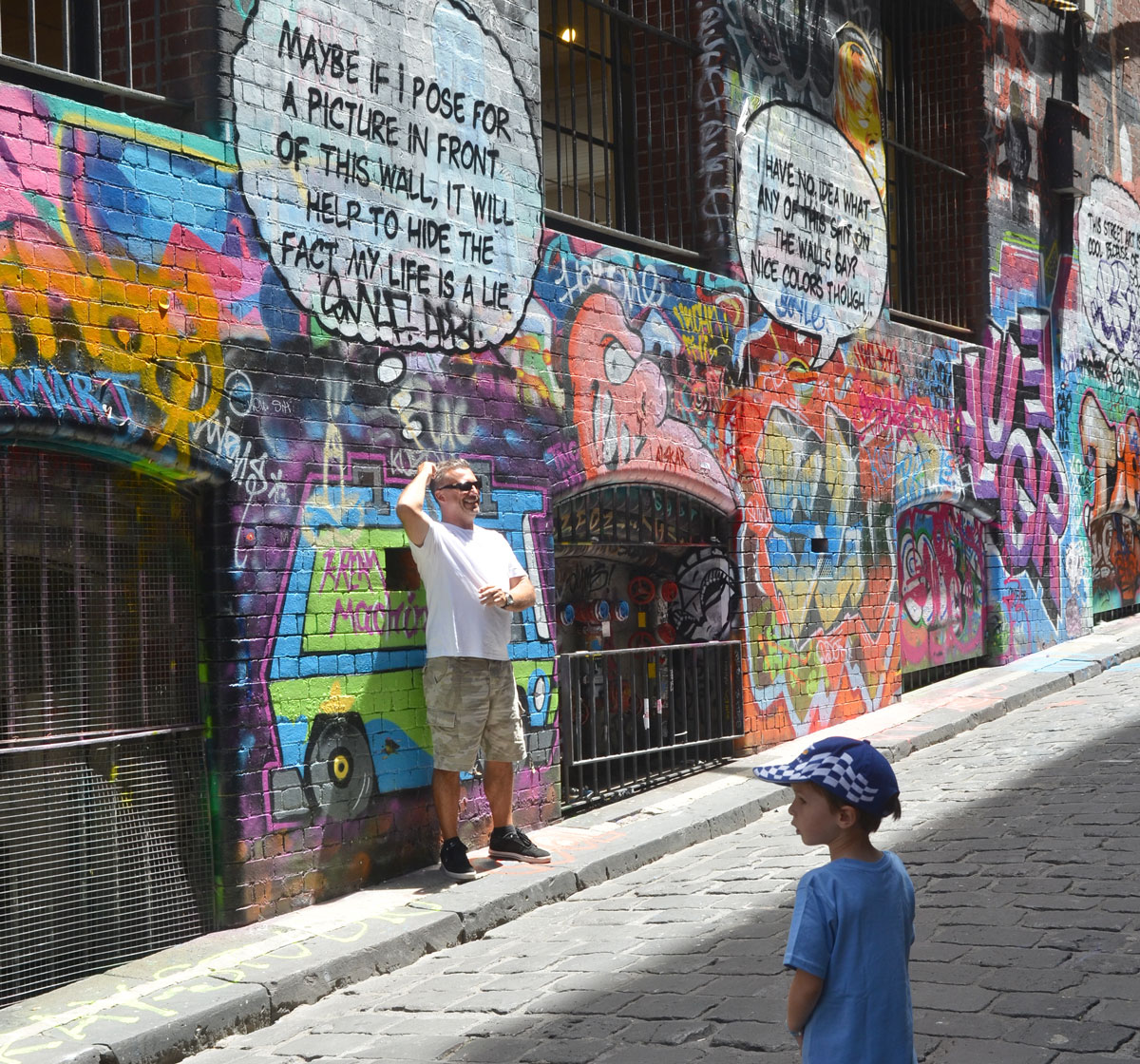 Lots of graffiti on the wall of Hosier Lane, windows with metal bars over them, a man poses in front of words that say "Maybe if I pose for a picture in front of this wall it will help to hide the fact that my life is a lie"