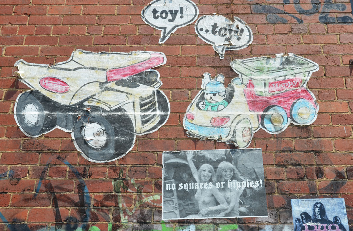 wheatpaste graffiti on a wall. A toy dump truck and a toy truck with a toy rabbut driving it. A poster of two topless women with the words not squares or hippies