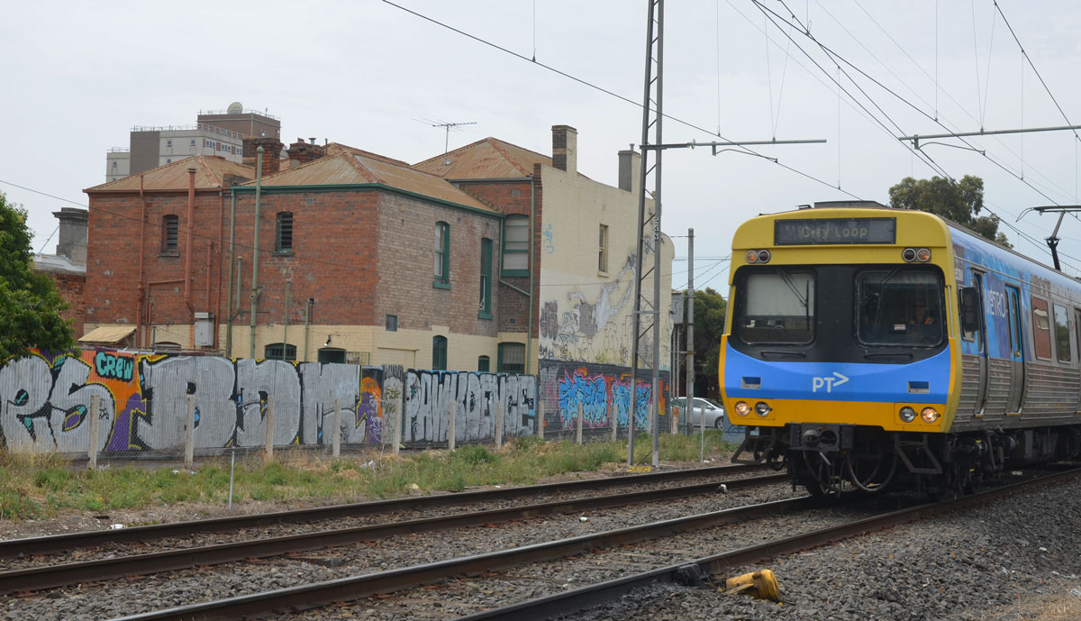 Melbourne PT train on train tracks as it passes by a wall of graffiti and some buildings behind the wall