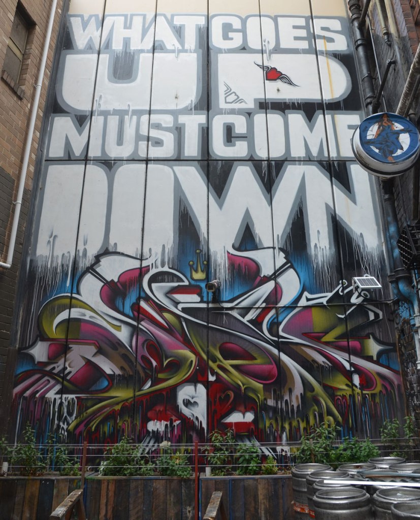 a large vertical mural at the end of an alley, with words written large that say What goes up must come down. Below the words is a swirly tag in red, white, and green