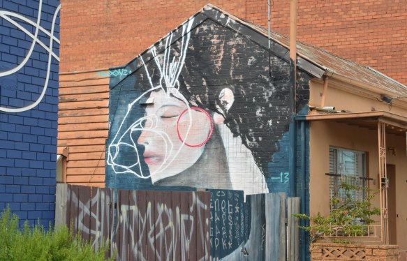 On the side of a small brown house is a large painting of a woman's head and neck. Short black hair, pink cheeks and her eyes are closed. The lower part of the picture is behind a fence. The picture takes up the whole side of the house. 