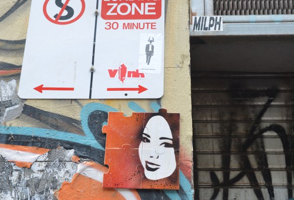 A small picture of a woman's face on a four piece puzzle on a wall in an alley, under a no parking sign