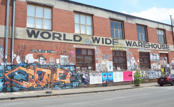 The side of World Wide Warehouse, a two storey brick bulding that is quite long. The lower level is cover with graffiti and street art except where the windows are