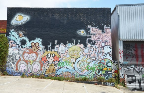 mural on a wall, the top is black sky, the bottom is a scene of people, a city and 2 flying saucers or UFO's. Not realistic looking, cartoonish