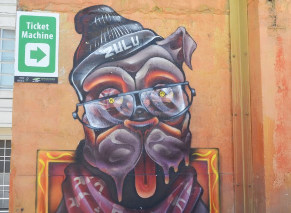 A dog's head. Drooling jaws, tongue sticking out, and wearing a hat with the name Zulu on it, on a wall in Rose Car Park 