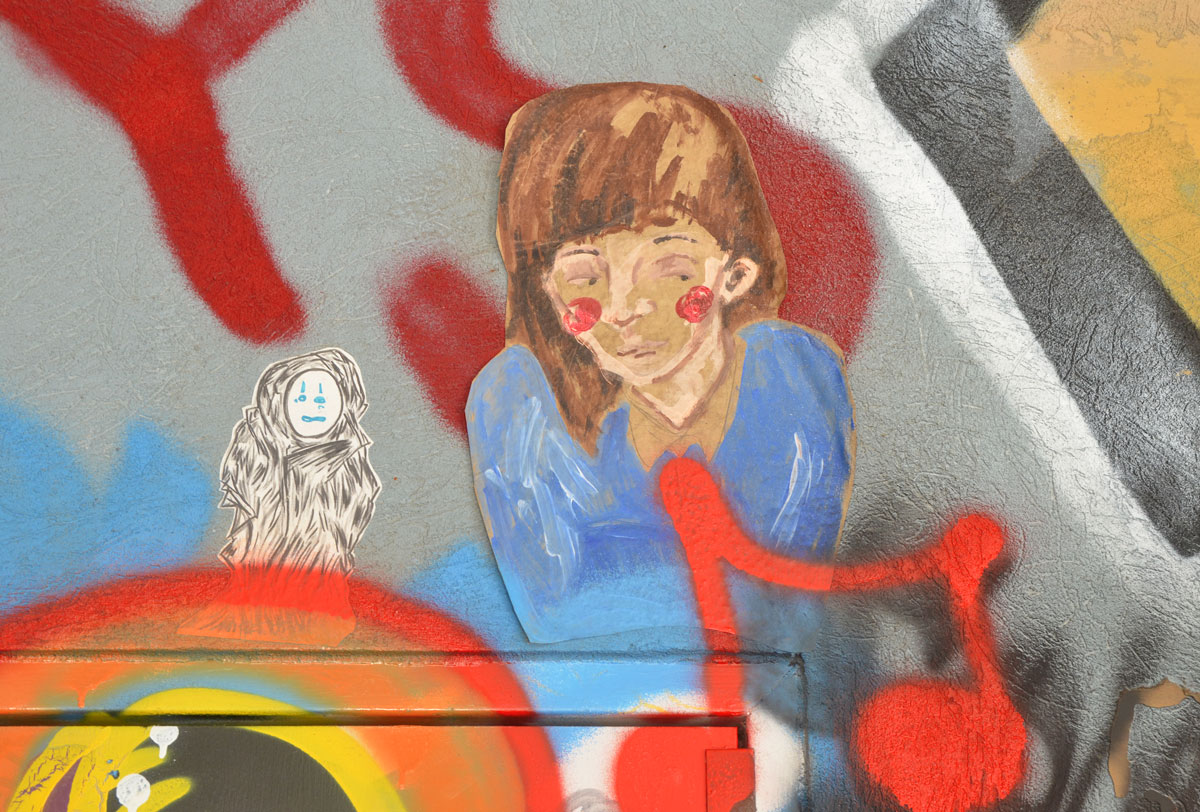 Two small paste ups. on the right is the head and shoulders of a girl with brown hair and blue top and on the left is a small black and white drawing of a little creature onto which someone has drawn blue eyes and mouth