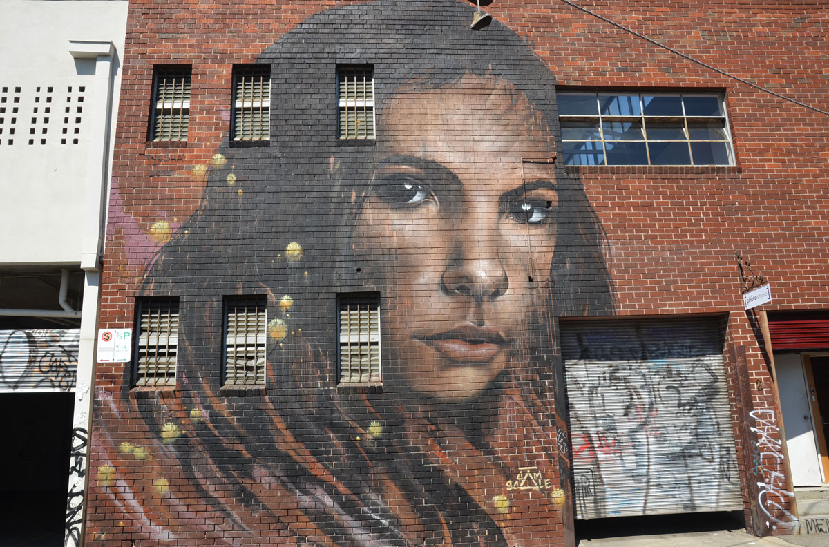 large two storey mural on a red brick building, by Cam Scale, of a woman's head and face, long black hair. 