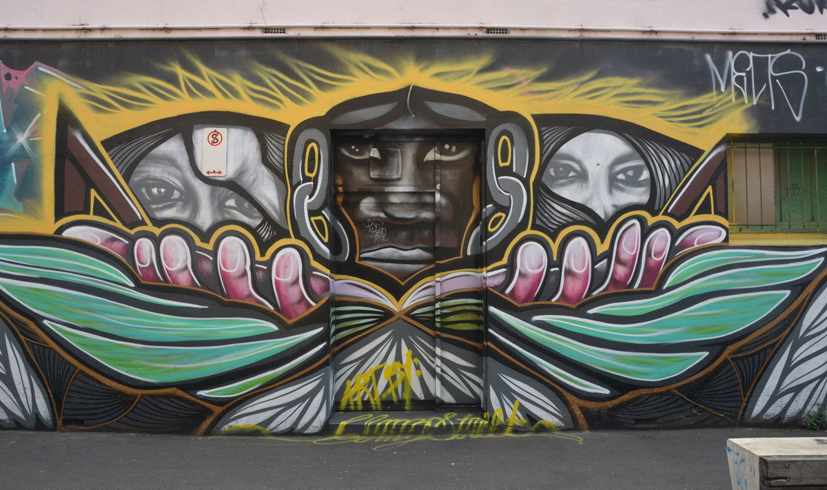 A horizontal mural on a wall with a door in the middle. A brown face on the door with two hands branching from it, each hand it holding a face. 