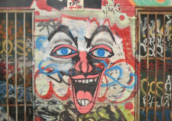 large clown like face painted on a wall between windows with metal bars. big laughing mouth and white face