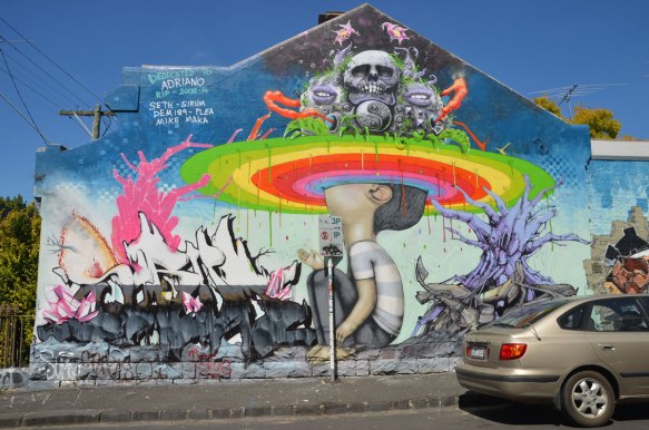 mural on Rochester steet in Fitzroy, boy sitting on ground with his head looking up and into a circular rainbow. Above him is a scene with skulls