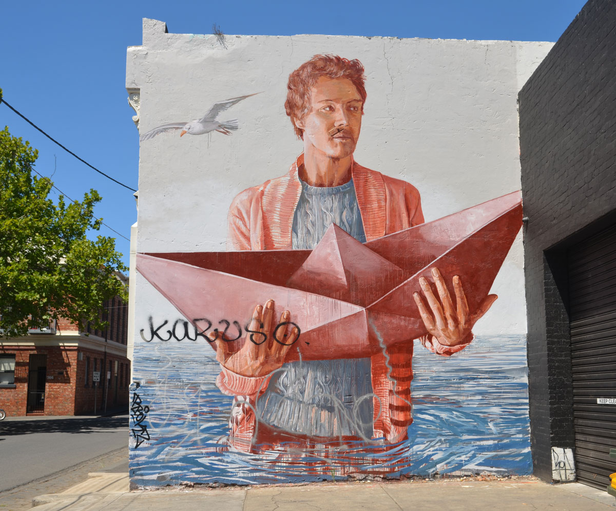 large mural of a man standing waist deep in water holding onto a folded paper boat in both hands. A seagull is flying past. The man is fully clothed. 