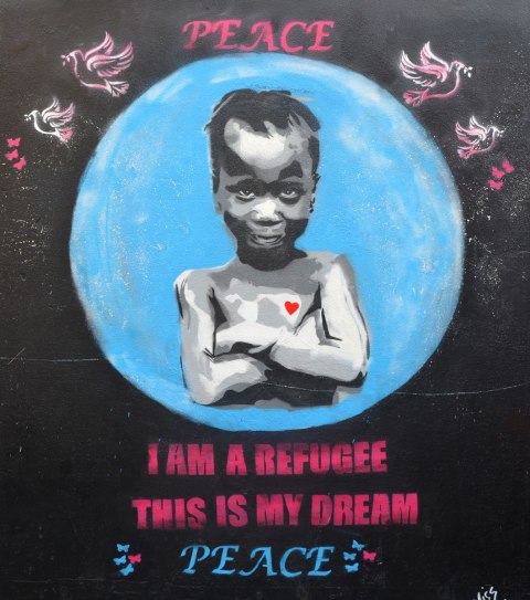 street art painting of a child with her amrs crossed, a smile on her face, and a red heart on her chest. Words "I am a refugee this is my dream. Peace"