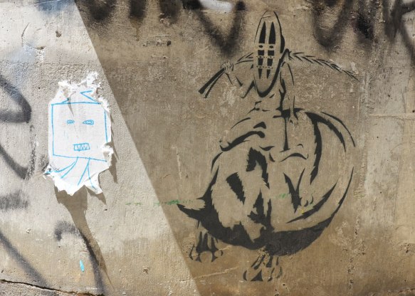 Two street art pieces. On the right is a black stencil of a person wearing an African mask and holding a branch behind his head. He is sitting on the back of an animal. on the left is a simplistic drawing of a square face with two little stick legs