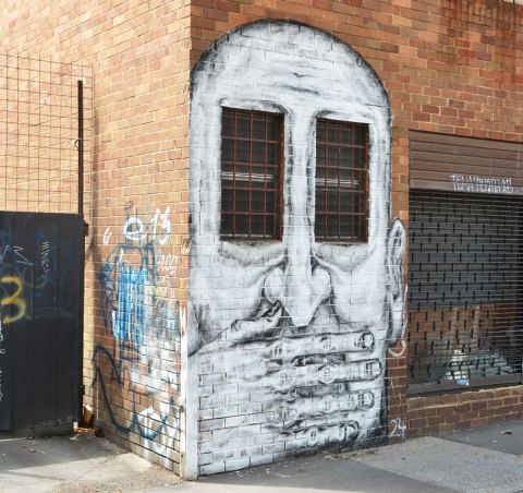 street art painting of a large white man's face with two windows as eyes, his hand is over his mouth