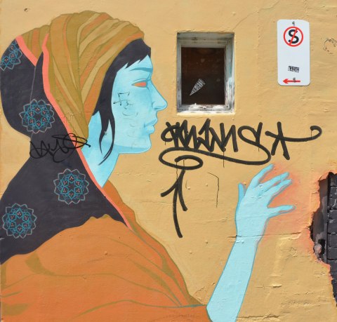 street art painting in Melbourne of a woman with light blue skin, an orange top and a two patterned head scarf, orange and gold stripes and black with blue and pink circular patterns on it. 