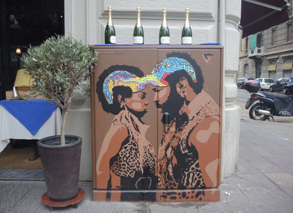 metal box on a sidewalk outside of a restaurant in Milan, painted brown with a picture of two black people, a man and boy, both with curly afro hair styles painted in rainbow colours. They are facing each other, nose to nose. 4 wine bottles are on top of the box. 