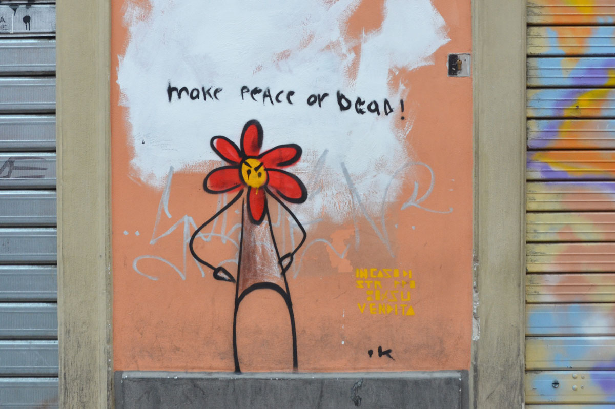 Small painting of a stick figure with its hands on its hips. Its face is a flower, looks like a red daisy with 6 petals. The words " make peace or dead are written above