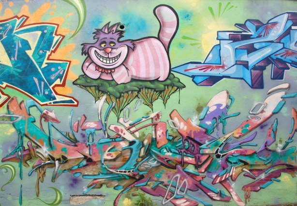 part of an Alice in Wonderland mural in a Montreal alley, looking down along the wall that the mural is on, a pink and white striped grinning cheshire cat sitting on a floating leaf.