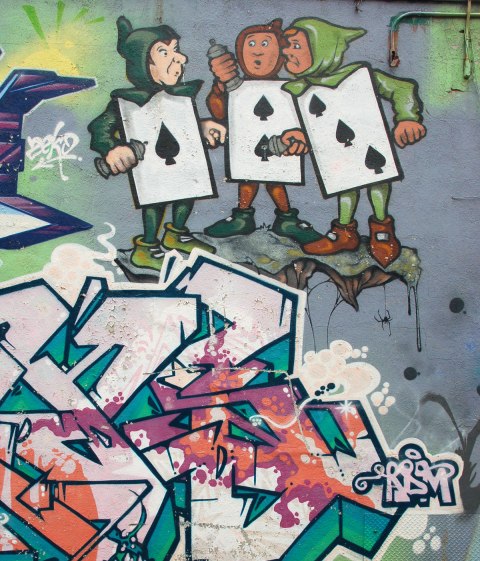 part of an Alice in Wonderland mural in a Montreal alley, looking down along the wall that the mural is on - three playing cards, the Ace, 2 and 3 of spades, as young men in black caps and tights, large cards for their bodies